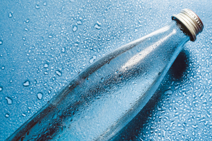 A glass bottle of water on blue background splashes drops of water on top. Quench thirst. Background water drops place for text copy space ACQUA