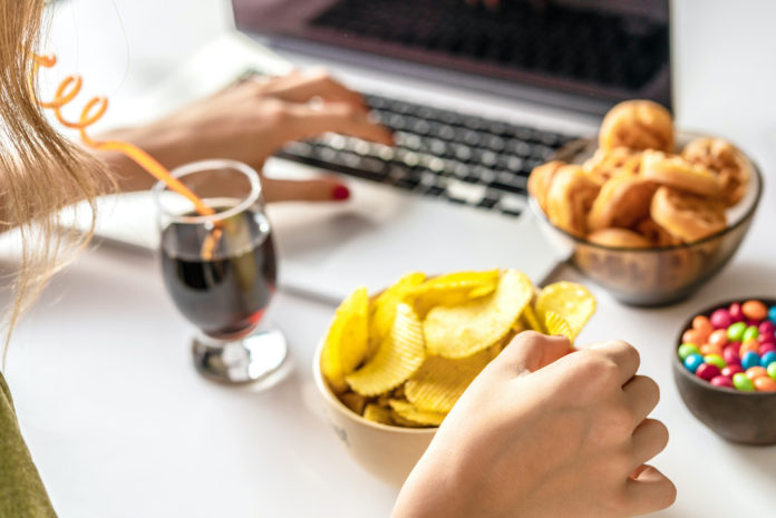 Girl works at a computer and eats fast food. Unhealthy food: chips, crackers, candy, waffles, cola. Junk food, concept. ALIMENTI ULTRA PROCESSATI