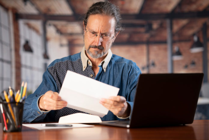 Angry stressed businessman opening envelope reading bad news in mail letter. Mad man feels frustrated about high bills, dismissal notice, bank debt, tax invoice or mistake sits at home office desk.