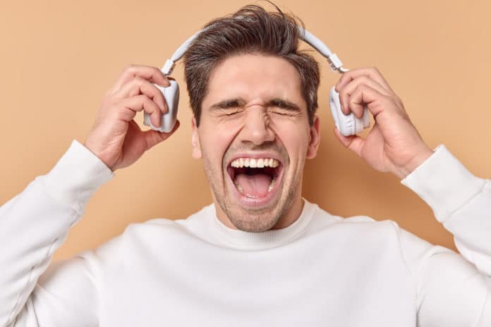 Emotional brunet European man exclaims loudly keeps mouth wide opened takes off stereo headphones listens music with high volume dressed in casual white jumper isolated over beige background. CUFFIE PER LA TV