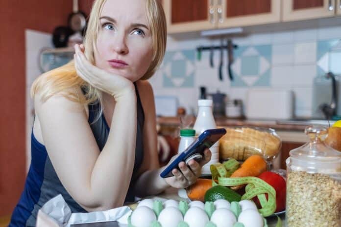 A woman checks the calorie content of food in a smartphone app. Calorie counting for weight loss. There are products for dieting on the table CONTA-CALORIE