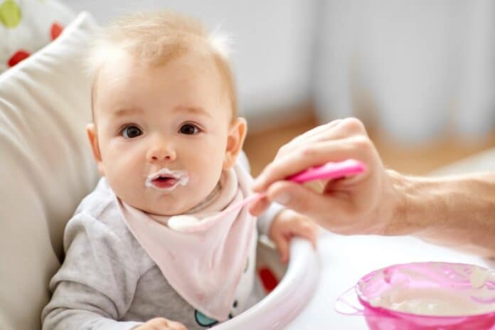 father feeding baby sitting in highchair at home ULTRAPROCESSATI