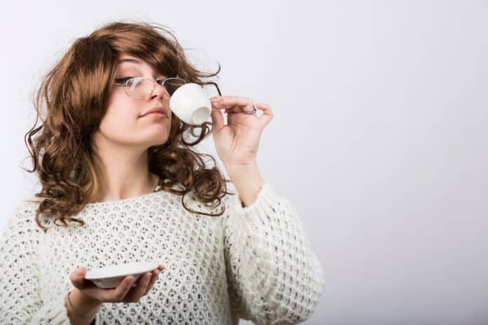 Signora con capelli mossi castani e maglione bianco beve il suo caffe da una tazzina , isolata su sfondo bianco CAFFÈ CAPSULE ESPRESSO