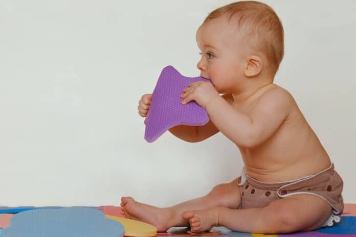 Cute baby biting foam puzzle shape is sitting on the colored mats. Active baby wearing cloth diaper. Isolated. Space for the text. Bpa