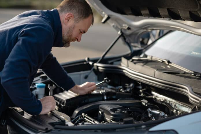 Man inspecting car engine with tools in hand, hood raised, focused on components, outdoor setting with natural light and blurred background